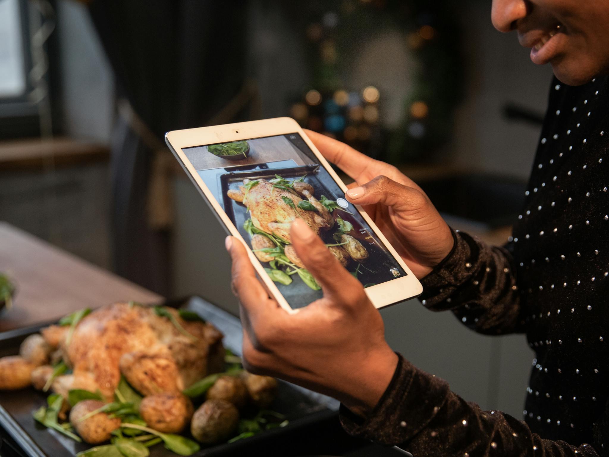 Person photographing a roasted chicken and potatoes with a tablet, highlighting food photography.